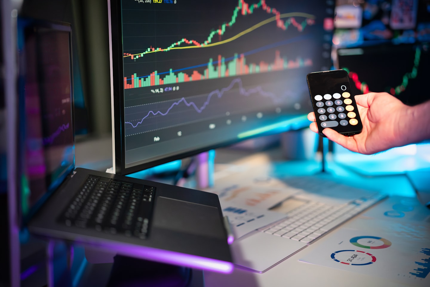 El, monitörlerdeki finansal verilerin yakınında hesap makinesi gösteren bir telefonu tutuyor.